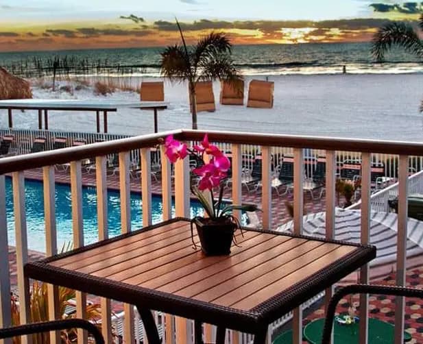 A balcony with a table and chair overlooking a pool and the ocean at sunset.